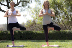 Two women stand on one leg on a mat with their hands pressed together in a mediative pose.