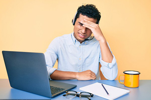 Image of a man in front of a computer grimacing and rubbing his forehead in annoyance