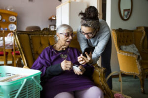 A young woman shows something on a phone to an elderly woman who is sitting down.