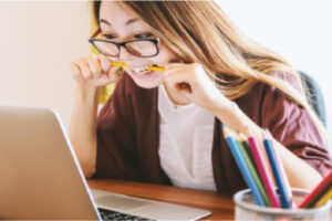 A woman with glasses stares at her computer and bites her pencil
