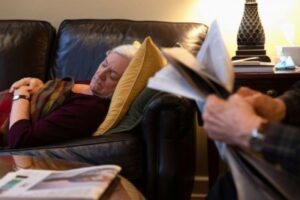 Image of an older female sleeping on a sofa and an older male reading a newspaper.