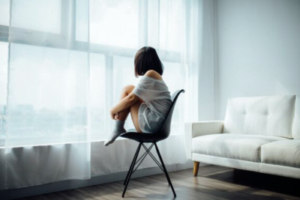 Woman sits on chair hugging their legs besides a window.