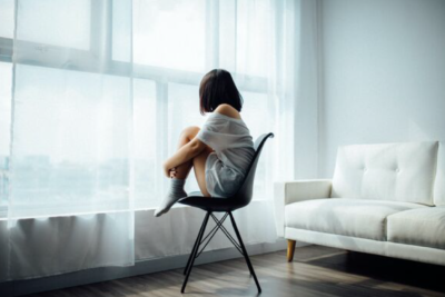 Woman sits on chair hugging their legs besides a window.