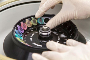 Close-up image of gloved hands removing a sample in a plastic vial sealed tube in a centrifuge.