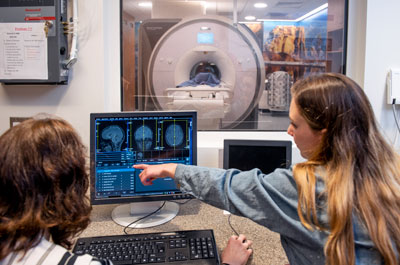 A woman sitting in front of an MRI room points at a brain scan on a computer screen