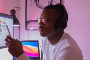 A man with glasses and headphone stares into his iPhone. In the background he leaves 2 open computers.