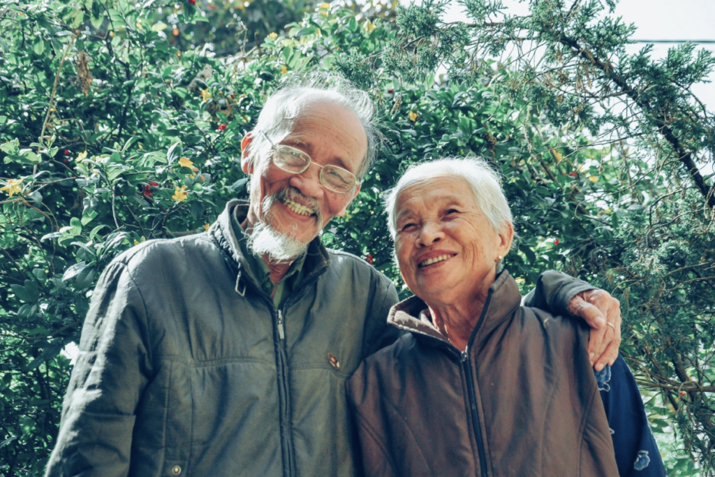 An elderly couple in jackets lean into each other, grinning at the camera. The person on the left wraps his arm around the other. Tall trees with red and yellow flowers fill the background