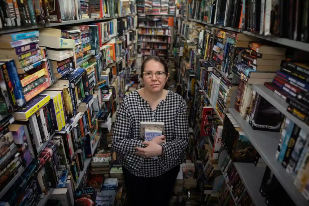 Rebecca Sharrock shoots a lopsided grin at the camera. She is holding two books but is surrounded by shelves full of books.