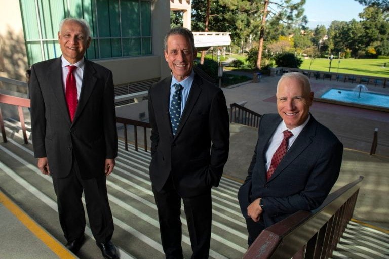 Three men in suits stand on a staircase outside and smile into the camera. A fountain runs behind them and further in the background is the lush green of Aldrich Park.