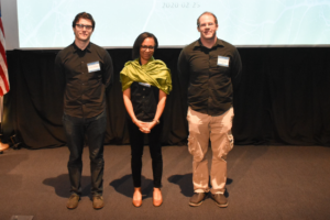 Three people stand in front of a projector screen and smile at the camera.