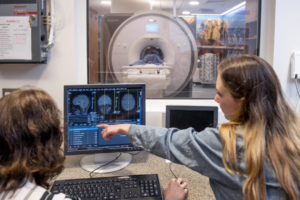 Two people observe a MRI scan through a glass window and looking at the brain scan on a computer screen. The woman on the right points at the scan.