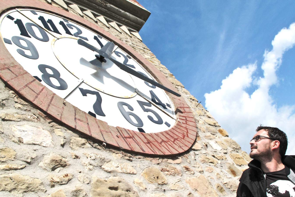 A man in the bottom right corner stares at a giant clock that takes over the entire left half of the image. In the background is a blue sky with some clouds