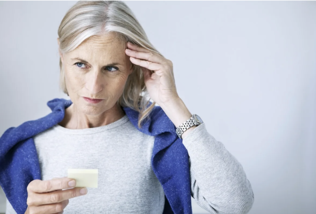 A stressed woman puts a hand to her head, likely trying to recall something while holding a sticky note in hand.