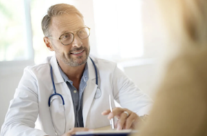 A doctor with glasses talks to a patient with a smile and pen in hand