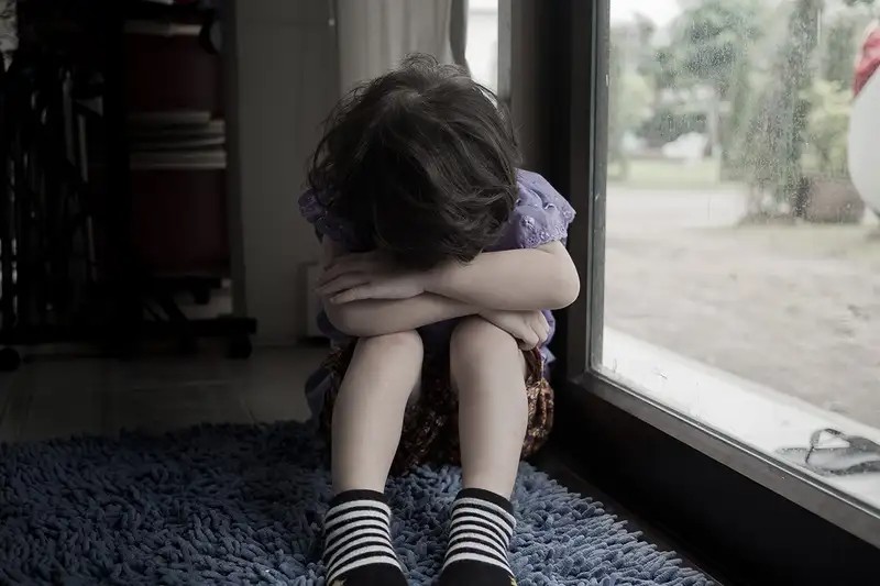 A child hugs their knees and hides their face in their arms by a window